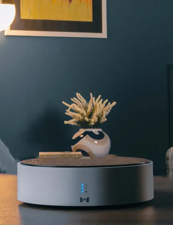 "MTronic MHub placed on a brown table with three blue vertical lights on and the MTronic logo in the bottom centre; a vase, blue walls and a yellow poster with white and black outline in the background.